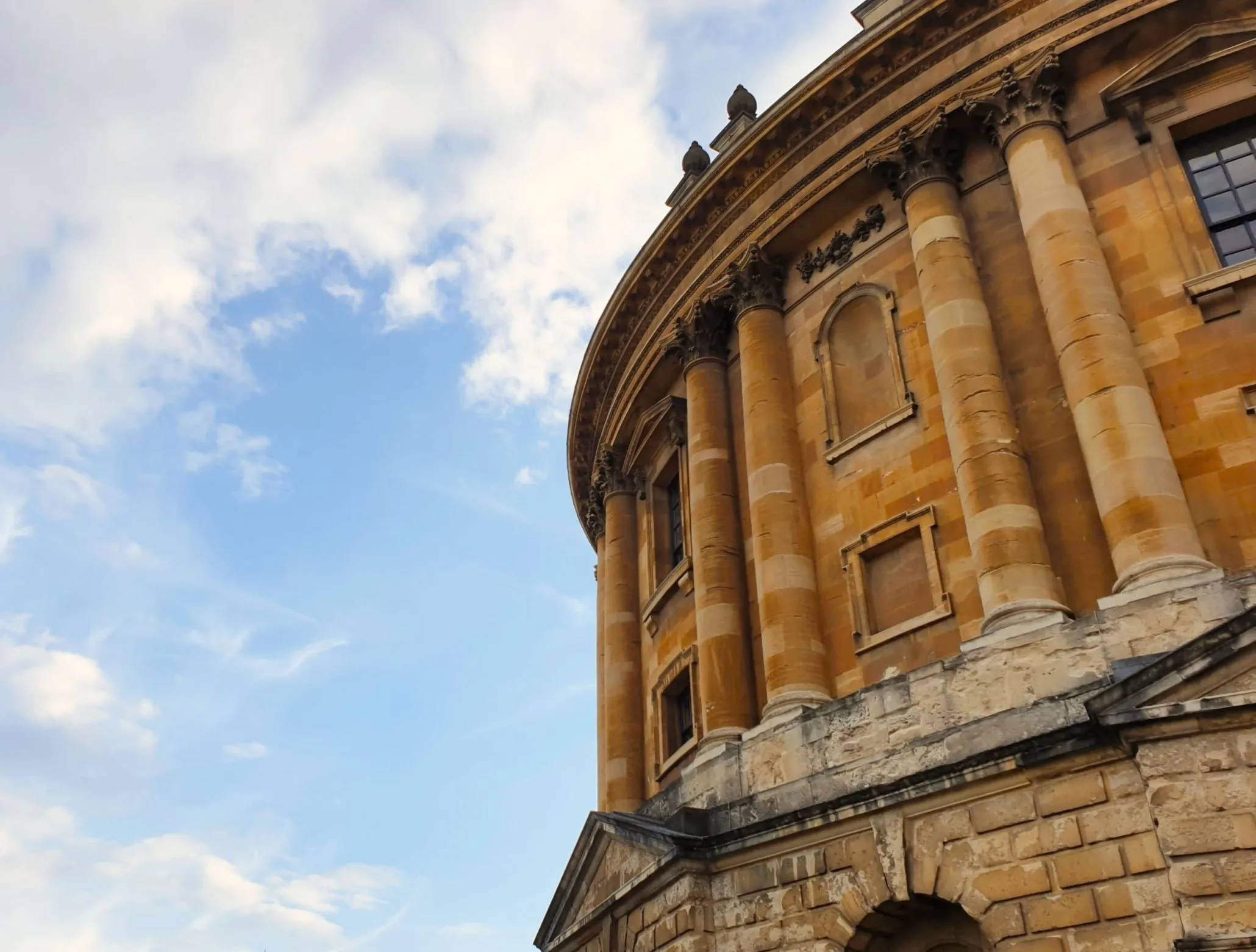 The outside of the Radcliffe Camera in Oxford. Behind, the sky is blue, with some clouds.